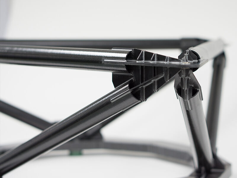 Image shows the completed TMT metrology fixture on a white surface and background. This detail shows a connection juncture, with four carbon fiber tubes bonded to a dark metallic and pointed fitting. Other tubes and the part of the circular base are in the background.